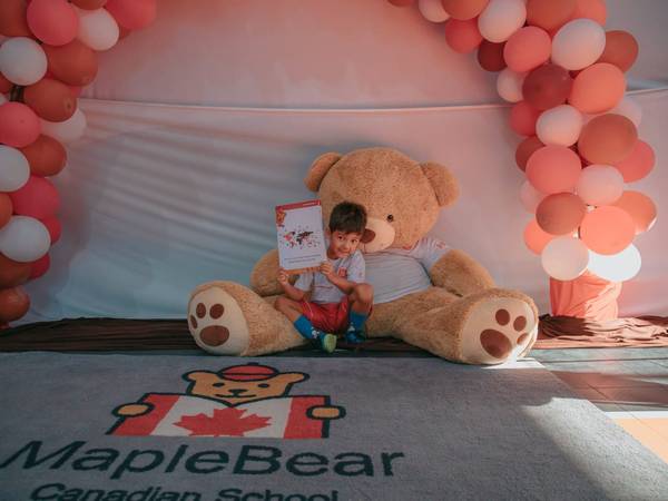 Urso de pelúcia da Maple Bear sob um arco de balões em evento da escola